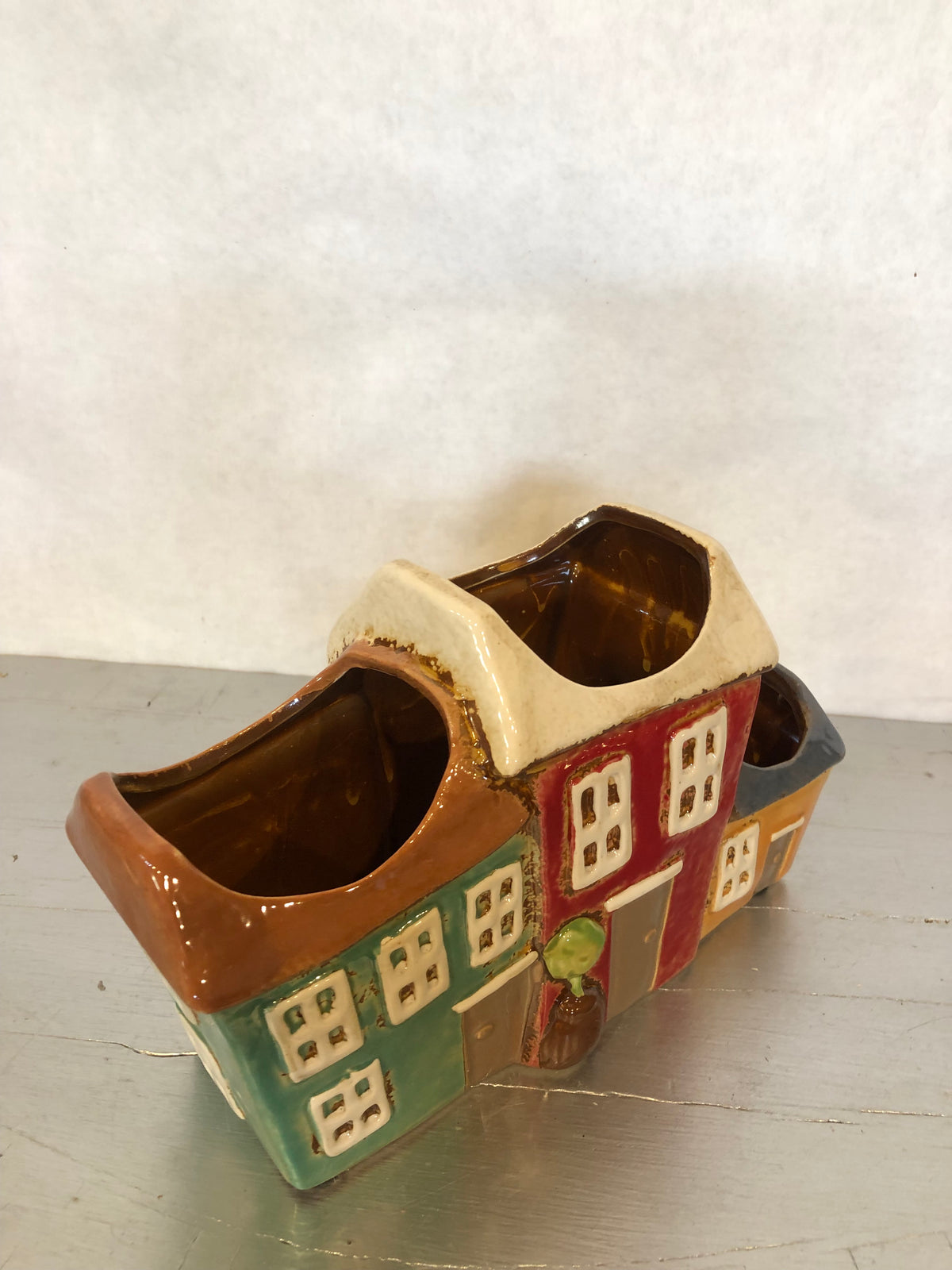 Vaso decorativo multiplo in ceramica smaltata a forma di casette colorate, dal design ispirato ai pittoreschi borghi mediterranei. Composto da tre vani porta piante integrati, è ideale per ospitare piccole piante grasse, cactus, erbe aromatiche, fiori secchi o composizioni decorative. Le vivaci tonalità verde, rosso e ocra, unite ai dettagli in rilievo di finestre, porte e alberello, lo rendono un elegante complemento d'arredo per soggiorno, cucina, ingresso, terrazza o ufficio. Perfetto per arredare ambien