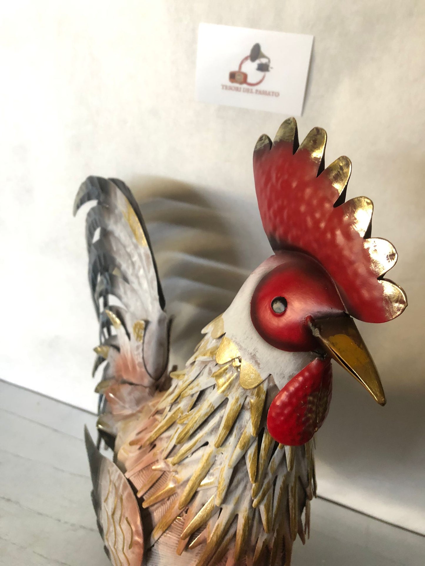 **Gallo decorativo in metallo dipinto a mano, ideale per arredare giardino, terrazzo, balcone, veranda o ambienti interni in stile rustico, country e shabby chic. Realizzato interamente in metallo con lavorazione tridimensionale delle piume, si distingue per la raffinata finitura nei toni del bianco, grigio, nero e oro anticato, impreziosita da cresta e bargigli rossi che donano un aspetto elegante e realistico. La struttura autoportante garantisce stabilità e consente di posizionarlo facilmente su qualsias