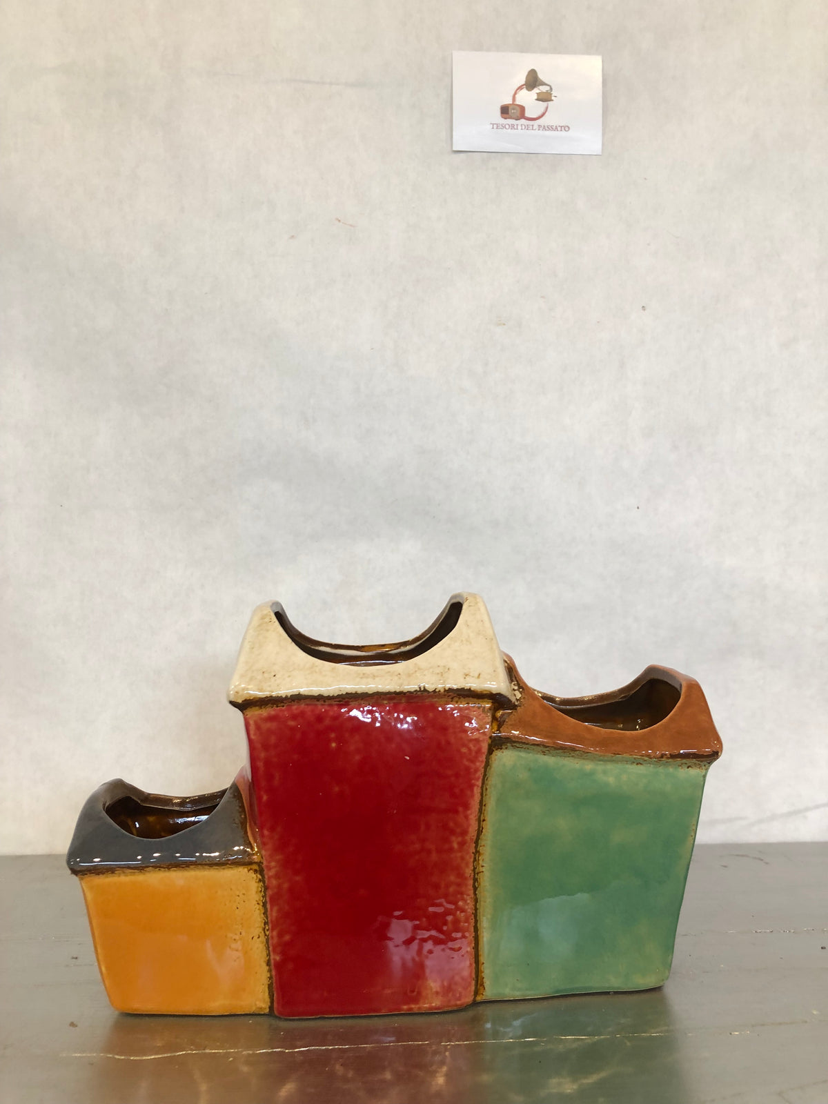 Vaso decorativo multiplo in ceramica smaltata a forma di casette colorate, dal design ispirato ai pittoreschi borghi mediterranei. Composto da tre vani porta piante integrati, è ideale per ospitare piccole piante grasse, cactus, erbe aromatiche, fiori secchi o composizioni decorative. Le vivaci tonalità verde, rosso e ocra, unite ai dettagli in rilievo di finestre, porte e alberello, lo rendono un elegante complemento d'arredo per soggiorno, cucina, ingresso, terrazza o ufficio. Perfetto per arredare ambien