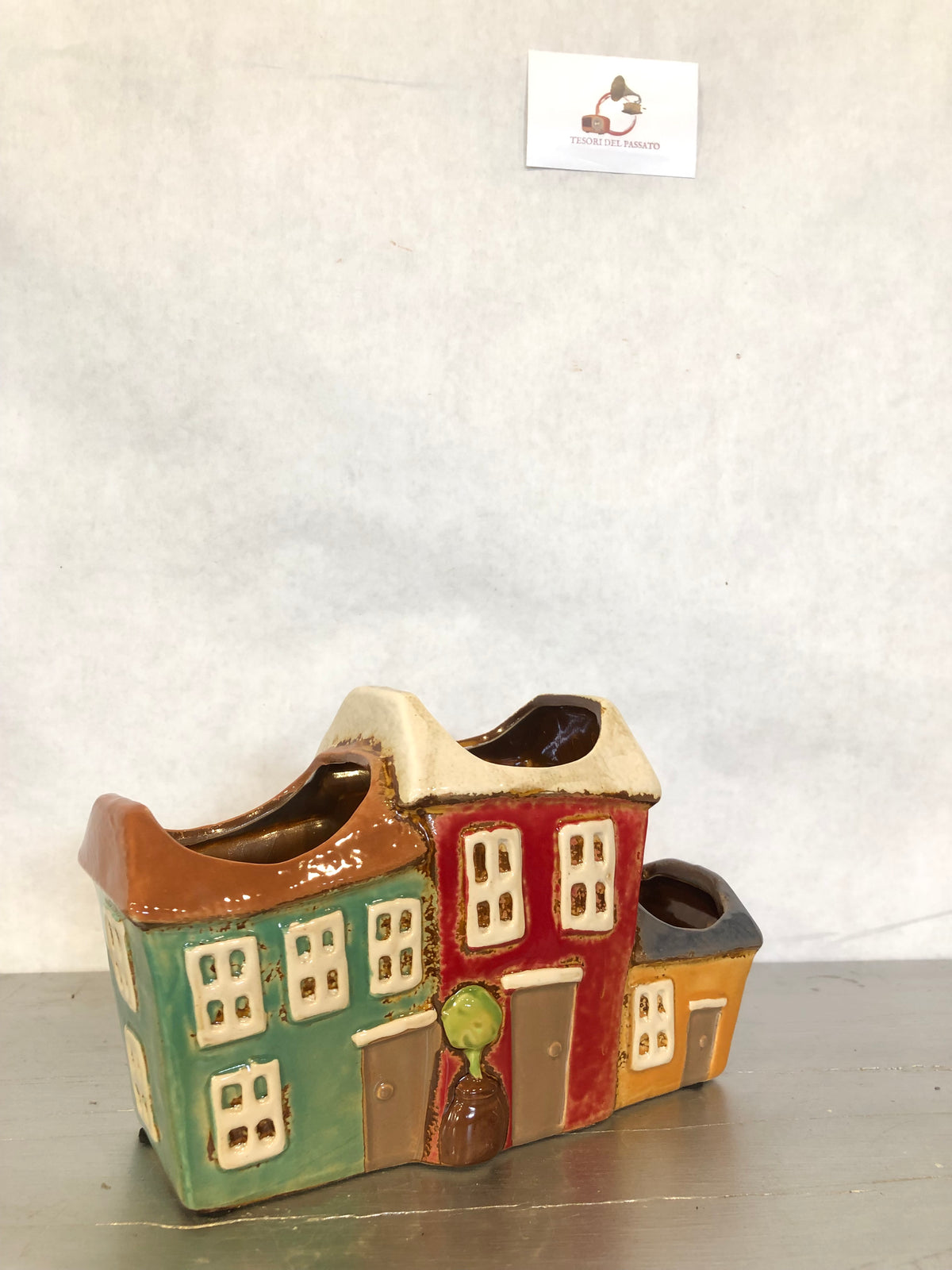 Vaso decorativo multiplo in ceramica smaltata a forma di casette colorate, dal design ispirato ai pittoreschi borghi mediterranei. Composto da tre vani porta piante integrati, è ideale per ospitare piccole piante grasse, cactus, erbe aromatiche, fiori secchi o composizioni decorative. Le vivaci tonalità verde, rosso e ocra, unite ai dettagli in rilievo di finestre, porte e alberello, lo rendono un elegante complemento d'arredo per soggiorno, cucina, ingresso, terrazza o ufficio. Perfetto per arredare ambien