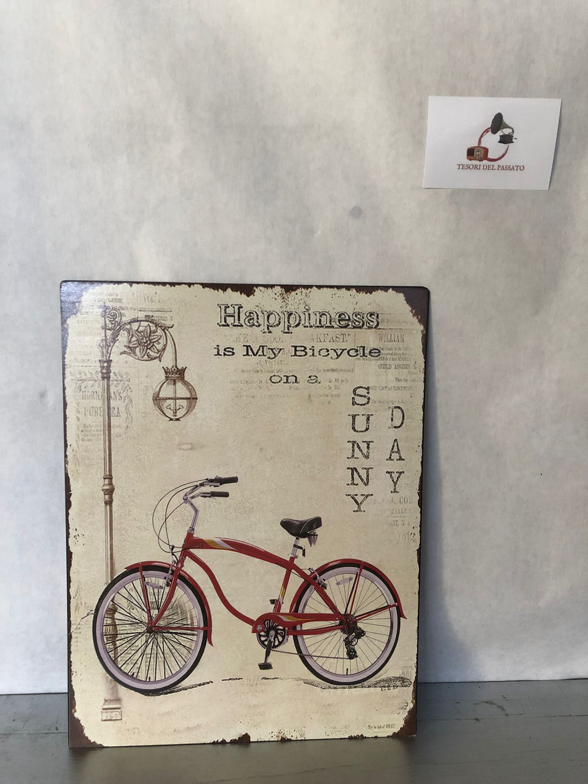 Quadro decorativo in stile vintage con elegante raffigurazione di una bicicletta retrò rossa accompagnata dalla celebre frase "Happiness is My Bicycle on a Sunny Day", perfetto per donare un'atmosfera rilassata e nostalgica agli ambienti. La grafica effetto anticato, impreziosita da dettagli come il lampione d'epoca e la finitura vissuta, richiama il fascino delle vecchie insegne decorative e dello stile industriale. Ideale per arredare soggiorno, ingresso, cucina, studio, ufficio, taverna, bar, caffetter