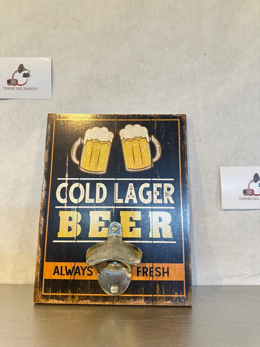 Apribottiglie da parete con stampa vintage "Cold Lager Beer", perfetto per arredare cucina, taverna, pub, bar, sala giochi, garage e angolo birra con un autentico stile retrò. Realizzato con robusta base in legno e apribottiglie in metallo fissato frontalmente, unisce funzionalità e design in un unico complemento d'arredo. La grafica effetto anticato raffigura due boccali di birra con la scritta "Cold Lager Beer – Always Fresh", richiamando le classiche insegne dei pub americani. Ideale per stappare comod